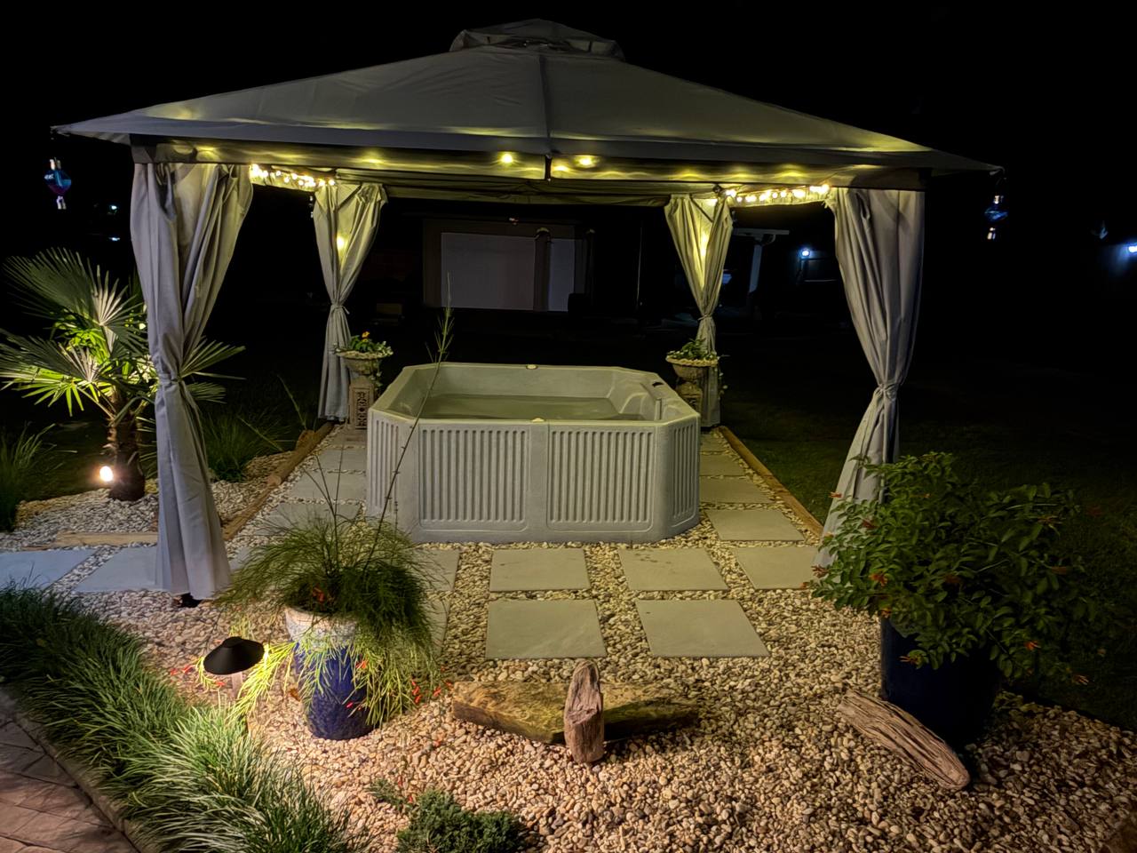 A hot tub beneath a curtained gazebo, ringed by uplit grasses and planters on a gravel and paver terrace at night