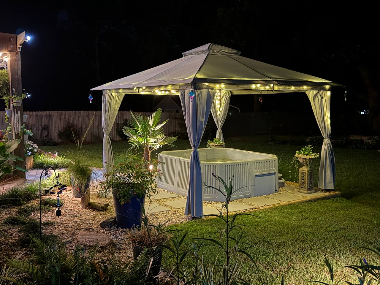 The lit gazebo seen across planting beds and young palms, with garden ornaments in the foreground