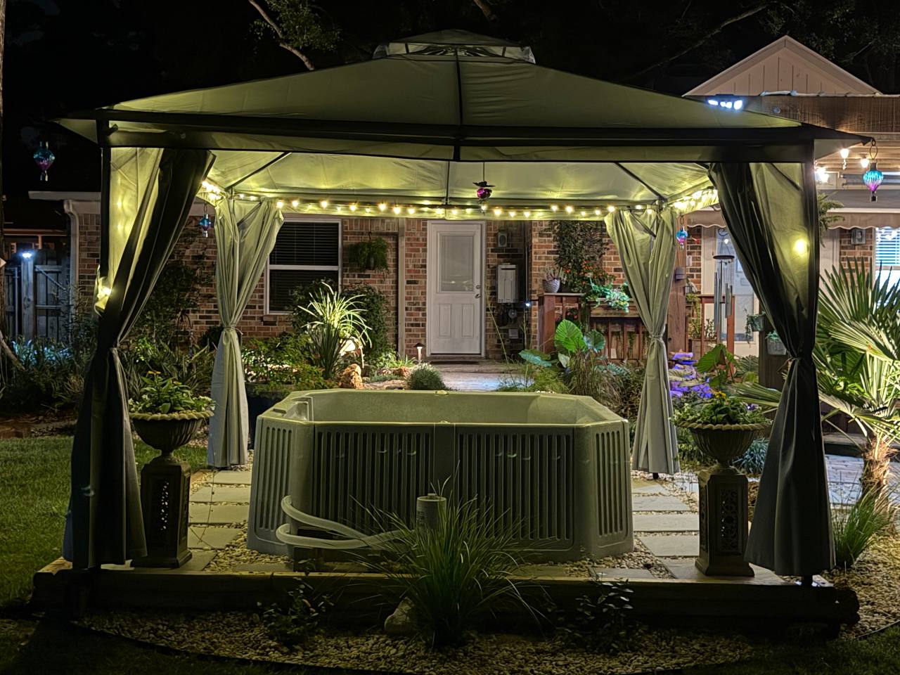 Straight-on view of the curtained gazebo and spa with the lit brick house behind it