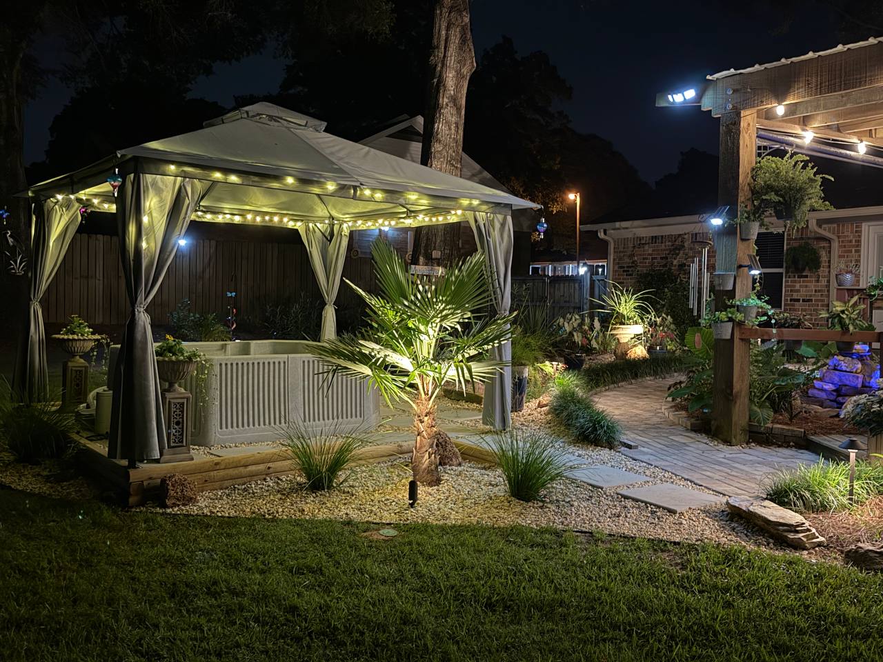 An uplit palm beside the gazebo, with a lit paver path running along a gravel bed
