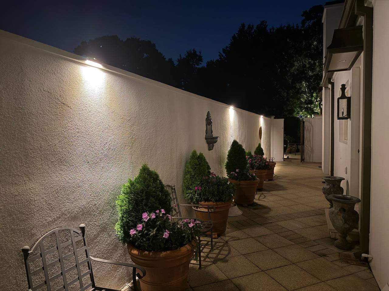 Long covered walkway along a garden wall with wall-mounted sconces, potted evergreens with pink flowers, and an urn fountain