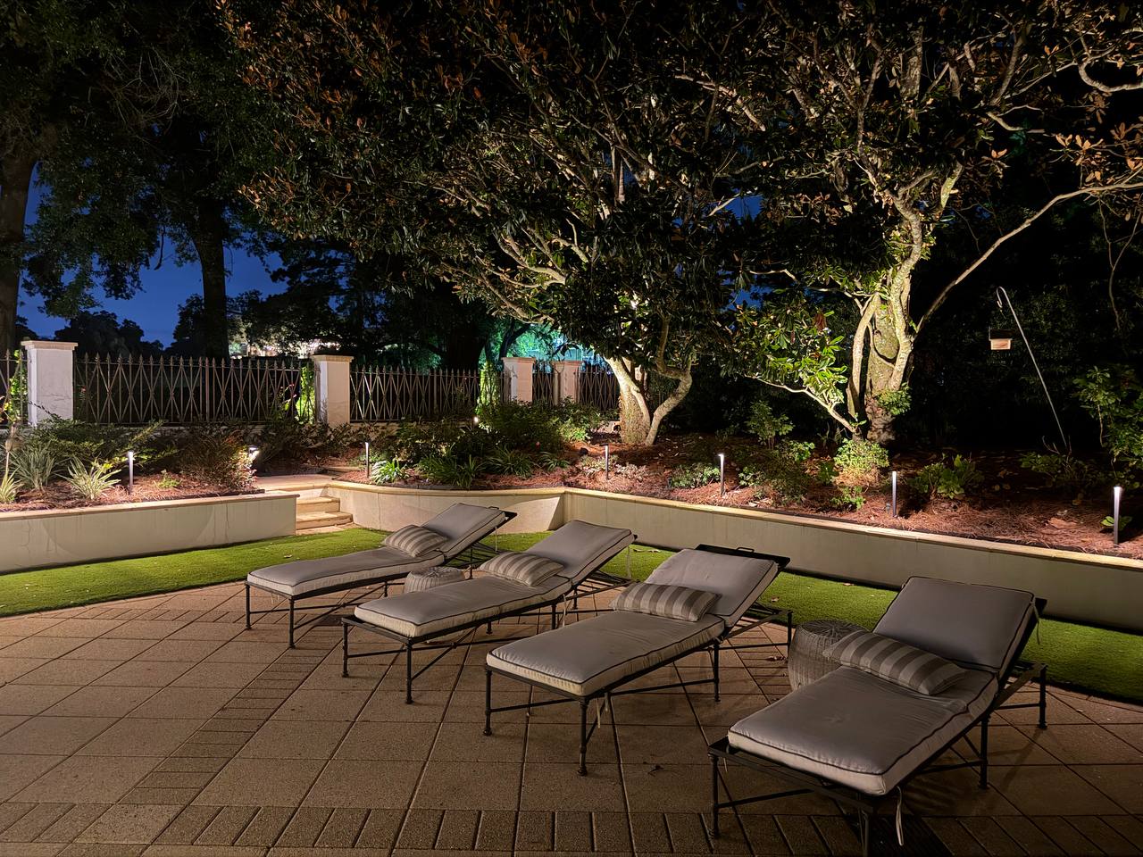 Four lounge chairs on a paver terrace beside a raised planting bed with path lights, beneath a wide live oak canopy and iron picket fence