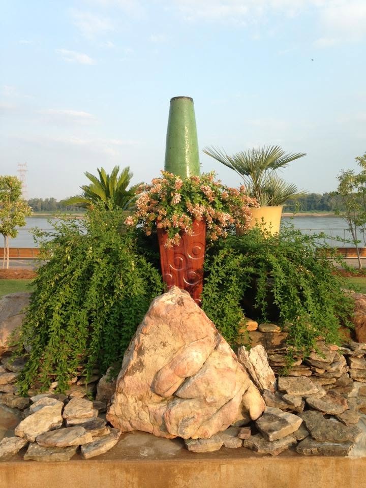 Natural rock water feature beside a waterfront rail with layered stone, ceramic urns, palms, and flowering plants
